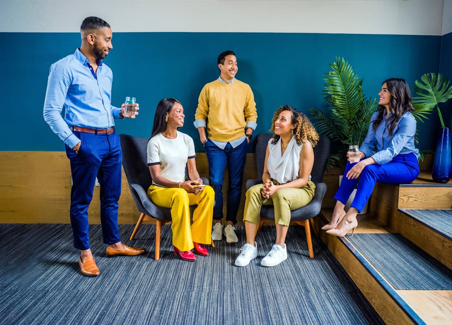 A diverse group of five professionals engaging in a casual indoor meeting, smiling and talking.