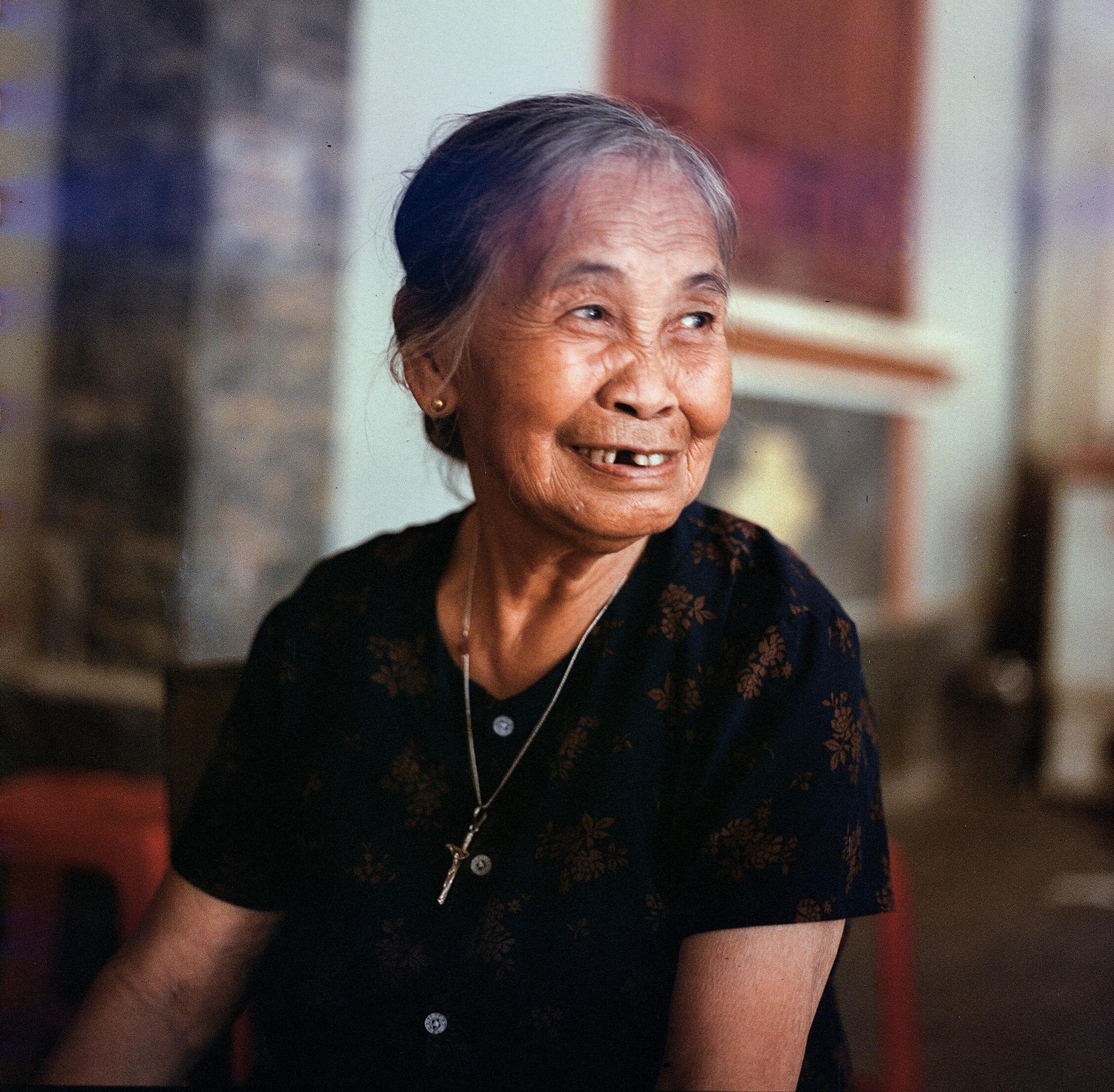 A smiling elderly woman with Asian descent sitting indoors, exuding warmth and contentment.