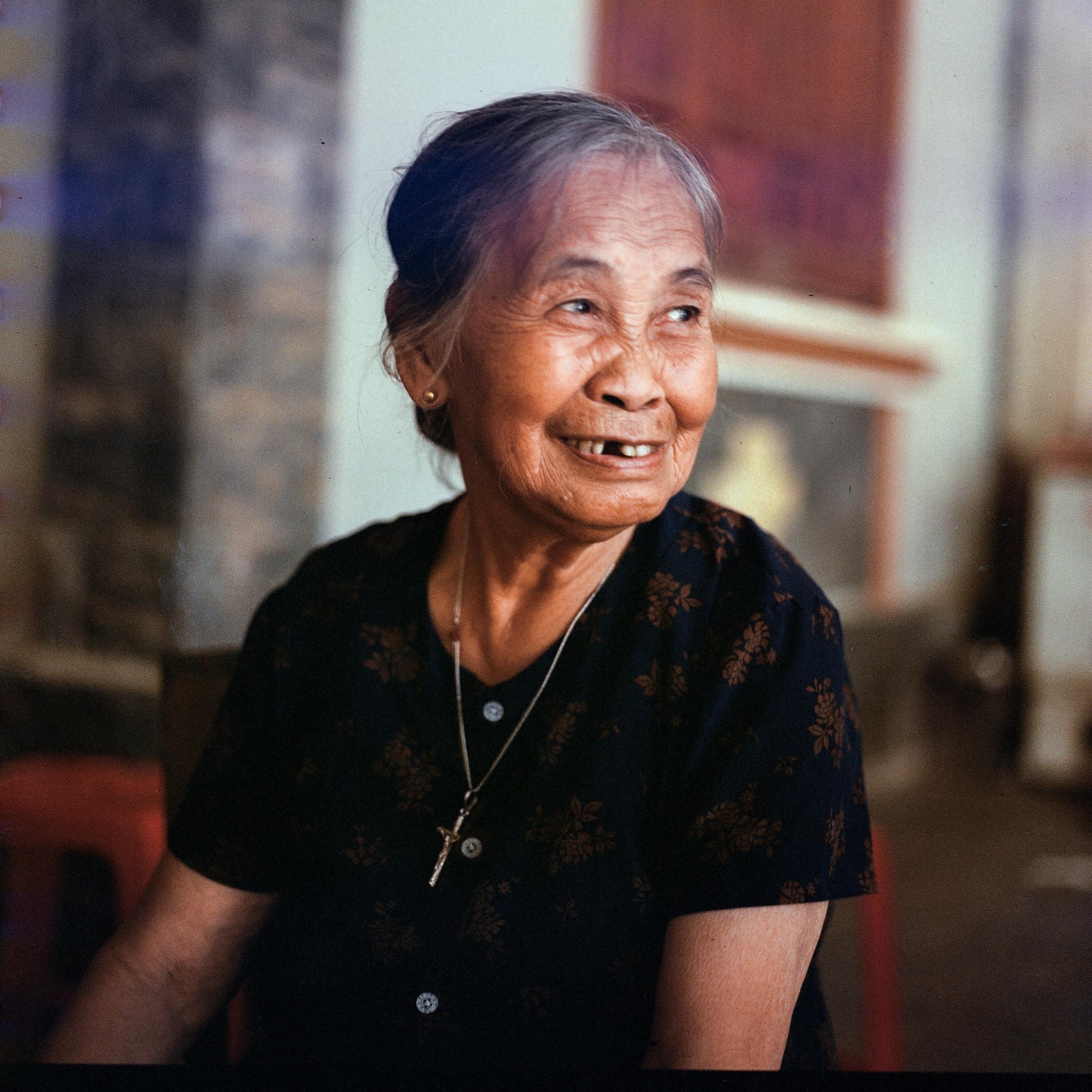 A smiling elderly woman with Asian descent sitting indoors, exuding warmth and contentment.